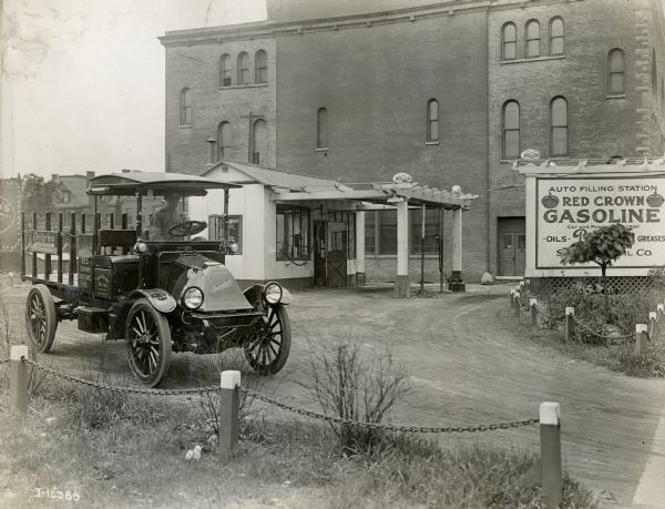 A man drives an International Model F (or 31) motor truck away from the Red Crown Gasoline auto-filling station. The truck was operated by Standard Oil and advertises many different types of gasolines, greases and oils for automobiles.