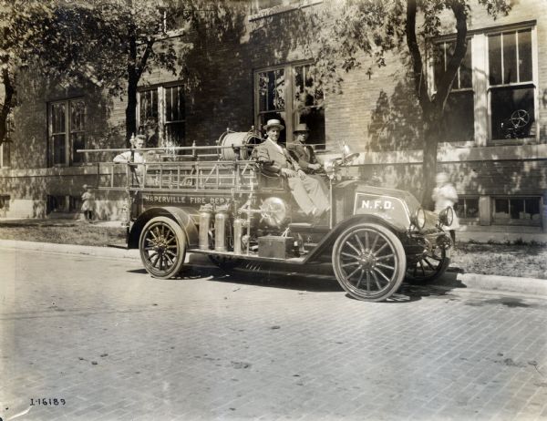 Three men on an International Model F 1916 fire truck operated by the Naperville fire deparment. Children are in the background.