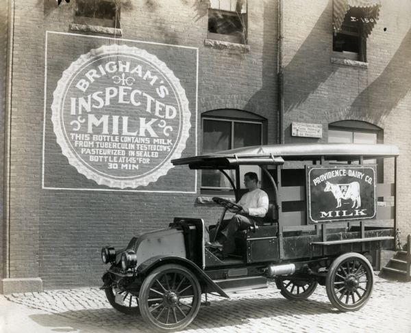A man sits at the wheel of an International Model F (or 31) truck operated by the Providence Dairy Company. The truck sits outside of a building with the sign "Brigham's Inspected Milk."