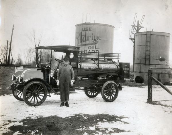 A man stands in front of an International Model F (or 31) truck advertising Standard Oil Company's Polarine, "Burn Perfect Oil." American flags are mounted to the front of the car and large oil storage tanks are in the background.