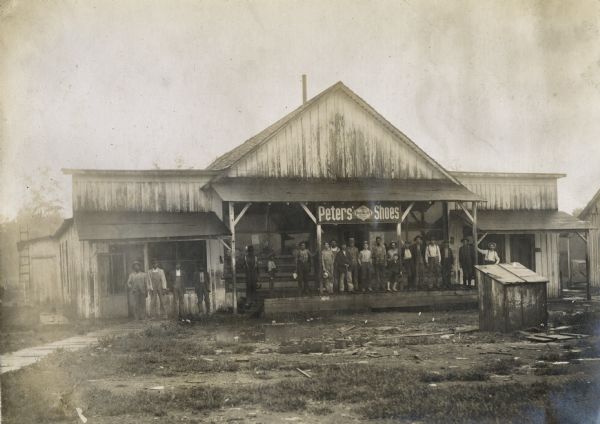 Men line up on the porch of "Peter's" shoe store. The store was likely at or near an International Harvester logging camp or sawmill.