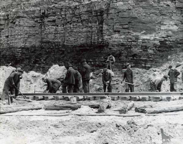 A group of men laying rails at Hawkins Mine while a man in the background is using surveying equipment near the base of a cliff face.