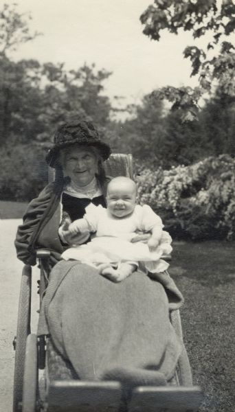 Nettie Fowler McCormick (1835-1923) holding her infant great-niece, Nancy Fowler Merle-Smith (b. 1921). Nettie is sitting in a wheelchair on a path, with shrubs in the background. Nettie and baby Nancy have similar delighted smiles.