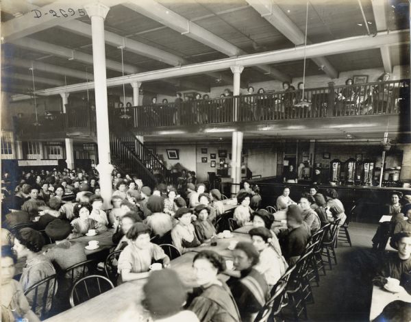 Female factory workers sitting at long tables with coffee cups at the Deering Twine Mill cafeteria. A large coffee bar is in the back right corner of the room.