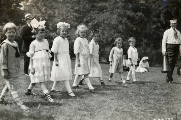 A group of children stand in a row while a man in an International Harvester hat stands beside them. The man is holding a megaphone, and the children may be preparing for a race at an International Harvester company picnic.