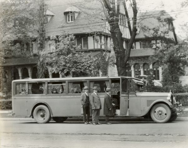 Three men stand on a residential street outside an International Harvester motor coach with "The Inter Cities Coach" written on the side. A large building in the background may be a boarding house. The bus was built on a model 52 or model 53 chassis.