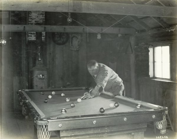 A man plays a game of pool in a cabin or shed. A wood-burning furnace is in the background.