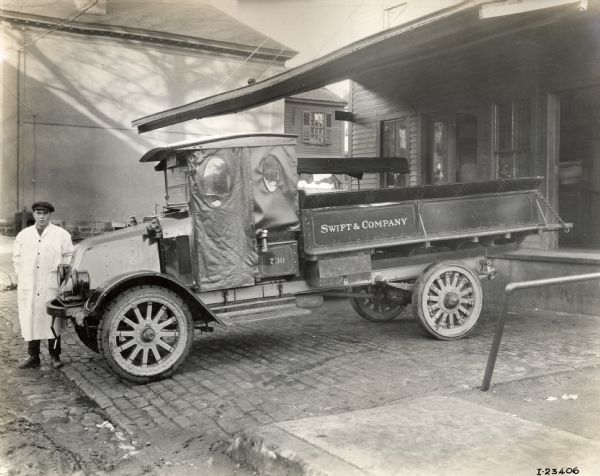 A man in a white smock stands near an International Harvester model G (or model 61) motor truck that is backed up to a loading dock at Swift & Company.