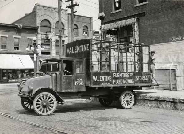 International Model G-61 truck operated by Valentine Furniture and Piano Moving and Storage. The truck is loaded with crates, possibly containing pianos, and is backed up to the side of a store. A man is standing at the back of the truck.