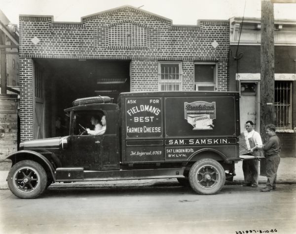 Two men are sitting in the cab of an International Model S truck used to deliver Fieldman's Best Farmer Cheese. Two men are loading packages into the back of the truck. The side of the truck has painted signs that read: "Ask For Fieldman's Best Farmer Cheese," "Sam. Samskin" and "Demand Breakstone's Downsville Cream Cheese." In the background is a brick building with a large, open garage door.