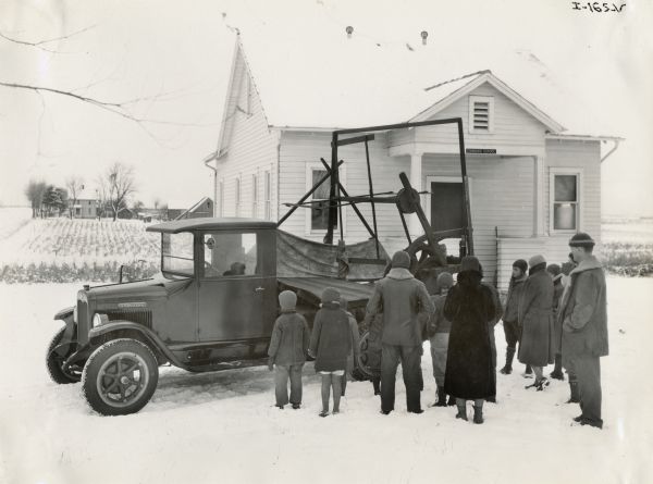 A group of children and adults gathers around an International truck carrying a replica of McCormick's original reaper. The reaper replicas were made for the "reaper centennial" celebration, an International Harvester promotional event. The truck is parked in front of a rural schoolhouse.