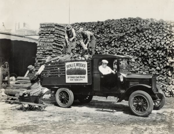 Men loading cut firewood from a large pile onto the back of an International Model S truck used by the Benj. E. Weeks Firewood Company.