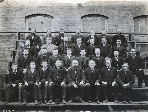 A group of factory workers sit on wooden benches in front of a brick building to have their portrait taken. The workers were employees of the McCormick Reaper Works, operated by the McCormick Harvesting Machine Company.