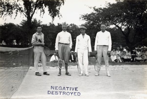 Four men holding racquets stand on a tennis court at an International Harvester Club picnic. Women are sitting in the background on the right.