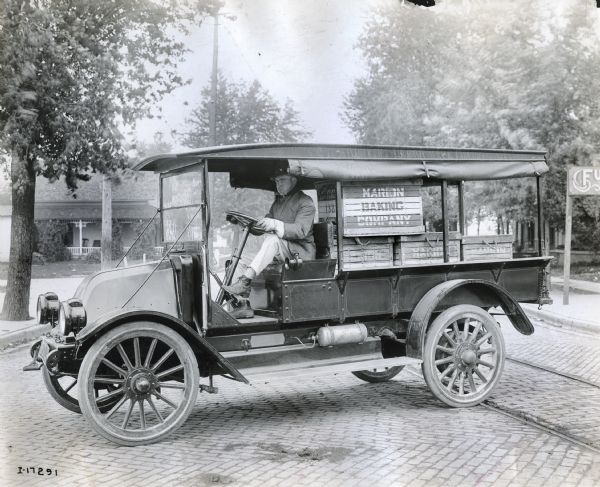 A driver sits behind the wheel of an International truck loaded with boxes from the Marion Baking Company. The truck is on a cobblestone street with cable car tracks, and may be a Model H or 21.