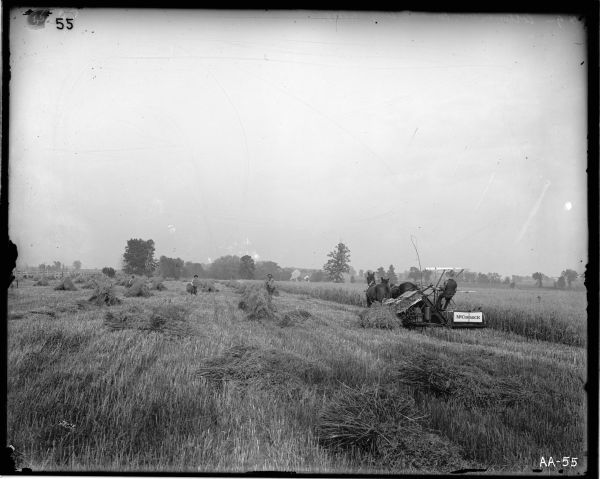 A farmer is operating a horse-drawn McCormick grain binder in a field, while workers are gathering sheaves of grain. The farmer is holding a raised switch.