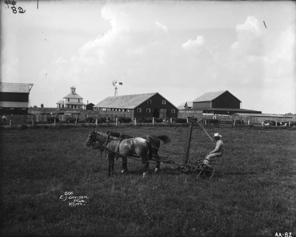 A farmer is sitting on a mower pulled by two horses in the middle of a field. Two other men appear to be herding cattle along a path beyond the field. A farm house, several farm buildings, and a windmill are also in the image. The initials "W.B.P.T" are painted along the top of a fence. "606/E.J.Davison/Photo./K.C.MO." appears to have been written on the photograph after it was developed.