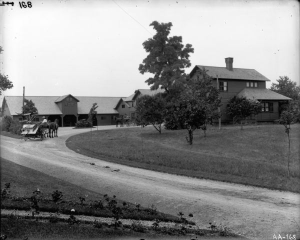 A man is driving a horse-drawn binder into the driveway of a farmstead. Another horse-drawn vehicle is partially visible in the driveway. This farmstead may be a model farm, an experimental farm of the late 19th and early 20th centuries.