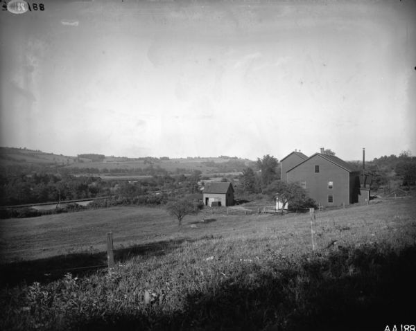 Rural scene of farmstead with a river or stream (?) in the background. The farm is bounded by a barbed wire fence, and there is a utility pole on the bank of the river.