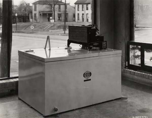 A six-can McCormick-Deering Milk Cooler on display in an International Harvester dealership.  A display case on the left near a window holds either toys or miniature models of agricultural machinery.