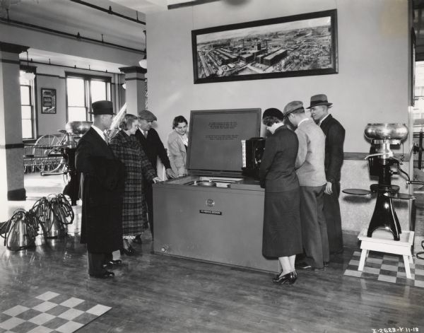 Three ladies and four men examine a McCormick-Deering 6-can milk cooler at a dealership or branch house. The photograph was taken in a large well-lit room with some other McCormick or milking related products. A large photograph of McCormick Works hangs above the cooler, and a poster advertising a McCormick All-Steel Spreader hangs in the back.