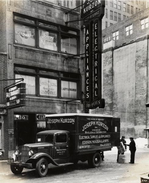 Two men load cargo into an International truck outside a Kurzon Appliances and Electrical Supplies building. Text on the truck reads: "Joseph Kurzon Inc. Wholesale Distributors Electrical Supplies Appliances/ Edison Mazda Lamps".