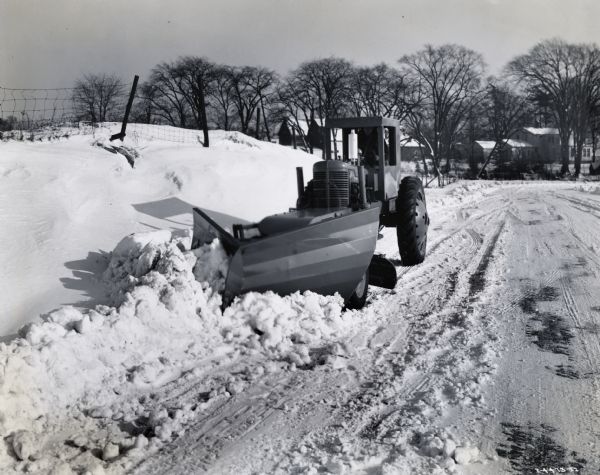 Man plowing snow with an International M tractor and snowplow. Original caption reads: "Then equipped with Trojan Vee-type snow plow as shown, the Trojan one-man speed patrol, which is powered by International M tractor, becomes a very efficient dual-purpose snow remover. Its regular blade supplements the snow clearing action when in down position and can be used to bench the snow on side bank when raised. It is shown at work near Batavia, New York, where snowfall has been unusually severe this season.  The Trojan Vee-plow with its expanding curve moldboard has a cutting width of 72 inches. It is raised and lowered hydraulically. The maintainer blade is 10 feet long and has a swing of 90 degrees. It has a side shift (per side) of 15 inches, clearance of 12 inches, and penetration of 5 inches. The Trojan maintainer is a fast-moving outfit, its speeds varying from 2 to 16 miles per hour, and of course on its pneumatic tires it can travel on all sorts of roads."