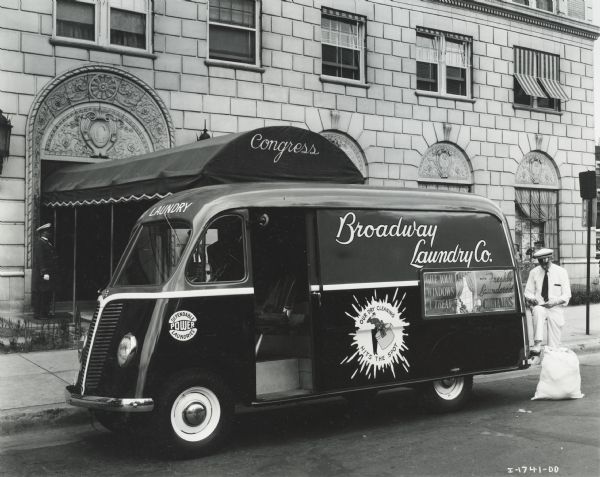 A man in a uniform standing at the back of an International Metro truck with one leg up on the bumper. He is writing on a pad held on his knee, and there is a bag of laundry on the ground next to him. The truck was owned by the Broadway Laundry Company. A building in the background has an awning labeled: "Congress", and a doorman in a uniform is standing at the entrance.