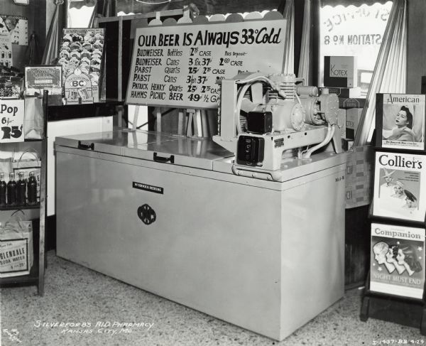 A cooler sits in Silverford's A.I.D. Pharmacy surrounded by advertisements, seeds, hygiene products, and magazines. A sign above the cooler reads "Our Beer is Always 33 degrees Cold" and lists the prices for various beers below. The original caption states: "McCormick-Deering 4 in line beer cooler." The three magazines on the right are the <i>American</i>, <i>Collier's</i>, and <i>Companion</i>.