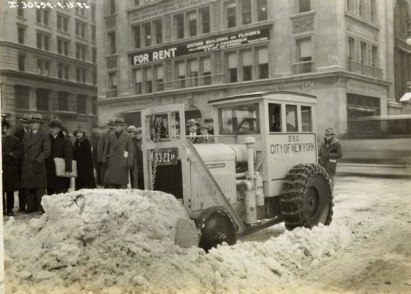 A man using a modified McCormick-Deering 10-20 tractor to clear snow from a city street. The cab of the tractor is labeled "D.S.C. City of New York."