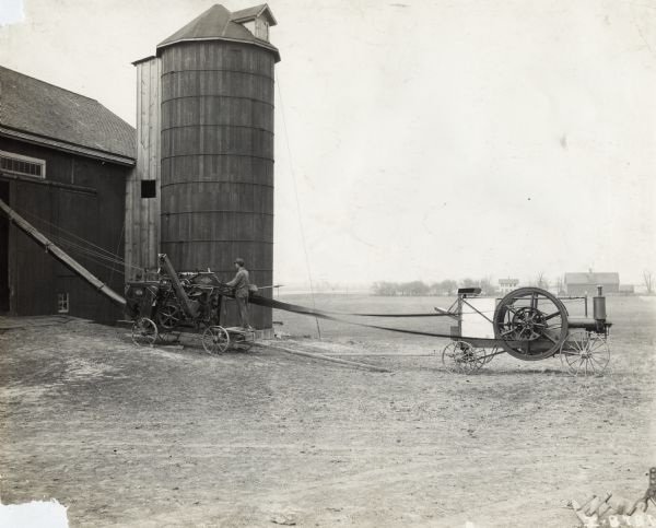 A man operating a Husker-Shredder near a barn and silo. The husker-shredder is powered with a belt connected to an engine.