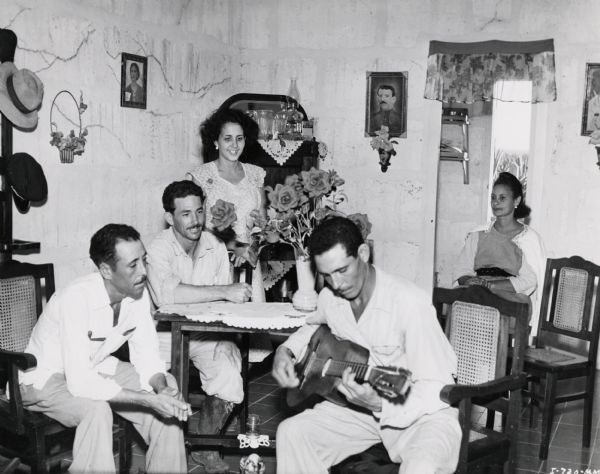 Three men and two women gather in a living room to listen to one of the men playing a guitar. The room is in the worker housing at an International Harvester sisal plantation in Cuba. Original caption reads: "Time off for recreation. This scene is of one of the native worker's living room."
