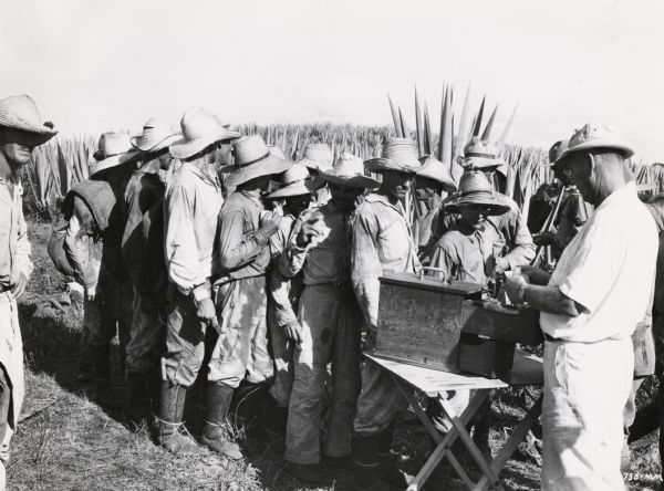 A line of workers forms in front of a table on an International Harvester sisal plantation in Cuba. The workers are waiting to collect their payroll checks. Original caption reads: "Payday on the IH(?) plantation. The third, fourth, and fifth men would appear to be counting up the take-home pay on their fingers."