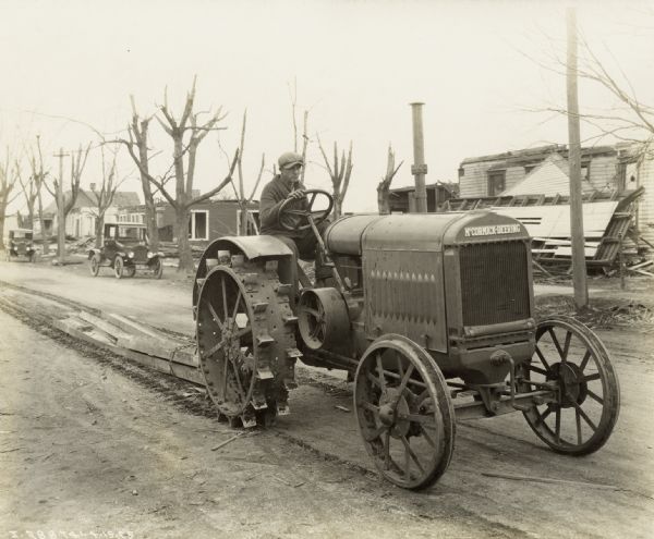 A man uses a McCormick-Deering relief tractor to drag metal pieces from a "tri-state tornado" damaged residential area. The trees along the street have broken branches, and damaged houses and debris can be seen in the background. The tractor may be a 10-20.