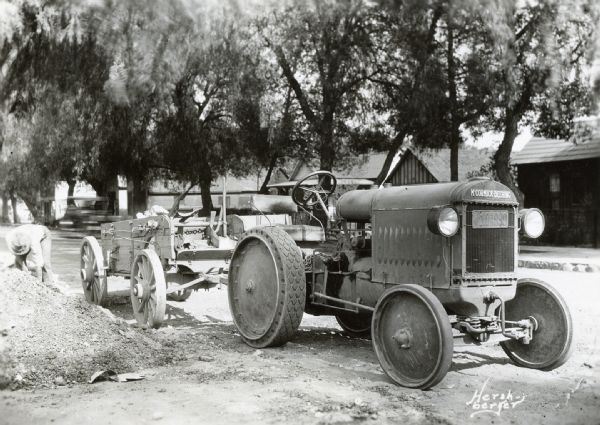 A man loads a pile of dirt or gravel into a wagon attached to a McCormick-Deering 10-20 industrial tractor. There are buildings in the background and an automobile (blurred by motion) driving by.