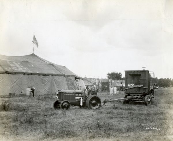 A man uses a McCormick-Deering 10-20 industrial tractor to pull a wagon at the Sells Floto circus. Children and adults are walking nearby, and a tent and large banners are in the background.