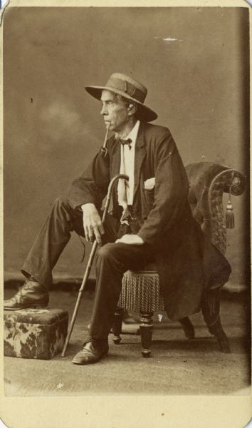 James Dorman Davidson (1810-1882) sits in profile in a plush chair for a studio portrait in front of a backdrop. He is wearing a wide hat, suit jacket, bow tie, and trousers. He smokes a pipe and holds a cane, and rests one foot on a low footstool.