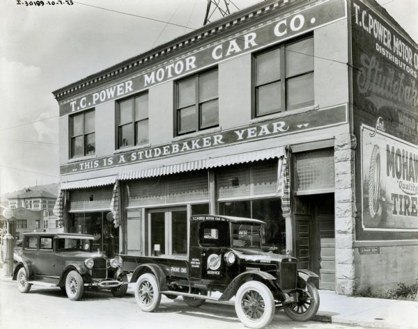 An International truck used by the T.C. Power Motor Company parked along the side of the road near the sidewalk outside the company office. There is a gasoline pump for Purol Gasoline on the left side. Other buildings and a hill can be seen in the background. The signs on the building read: "This is a Studebaker Year" and "Mohawk Tires."  The lettering on the truck reads: "Helena, Great Falls, Billings" and "Studebaker Service." There is a sign inside the windshield of the first car that says: Montana State Fair / (obscured) September 7-8-9-10."
