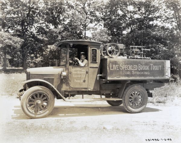 A boy sits behind the wheel of an International truck used by the Pepperridge Lake Trout Hatchery. The truck is parked along a dirt road in a wooded area and is carrying a small stationary engine. Text on the truck reads: "Live Speckled Brook Trout for Stocking."
