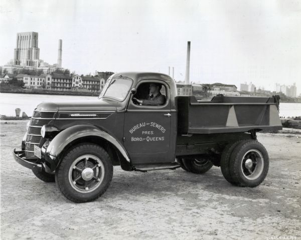 A man parking an International D-30 dump truck used by the Bureau of Sewers near a river or harbor. On the opposite shore in the background are buildings, skyscrapers, and smokestacks.