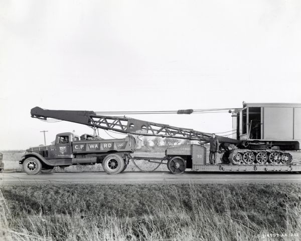 View from side of road of an International Model A-8 truck marked "C.P. Ward, Inc." hauling a trailer with a large crane. One man is driving the truck, another man is standing on the trailer near the crane. In the background is a field with a house and trees.