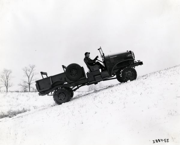 An International truck used by the U.S. Marine Corps is driven up a snow-covered hill by a man wearing a jacket, gloves, and baseball cap. The original caption reads: "Springfield Works, M-2-4-233, U.S. Marine Corps."