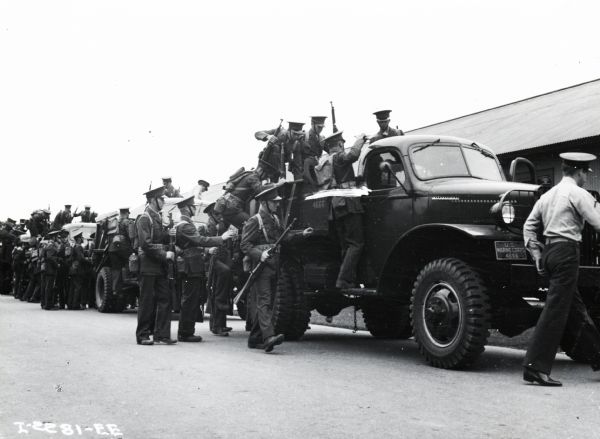 Groups of uniformed Marines and carrying rifles are climbing into the back of a number of International trucks. There is a long building in the background. The original caption reads: "Group of Marines shown in Photograph I-2267-EE loading into International trucks for transport to rifle range. Truck in foreground is Model M-3-4 4-wheel-drive with standard cab."