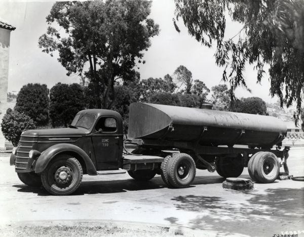 An International truck outfitted with a fuel oil tank parked in a lot beneath trees. There is a man working in back of the truck, and buildings are in the background. The original caption reads: "Model D-60 with semi-trailer tank used to transport fuel oil at Marine Corps Base, San Diego, Cal."