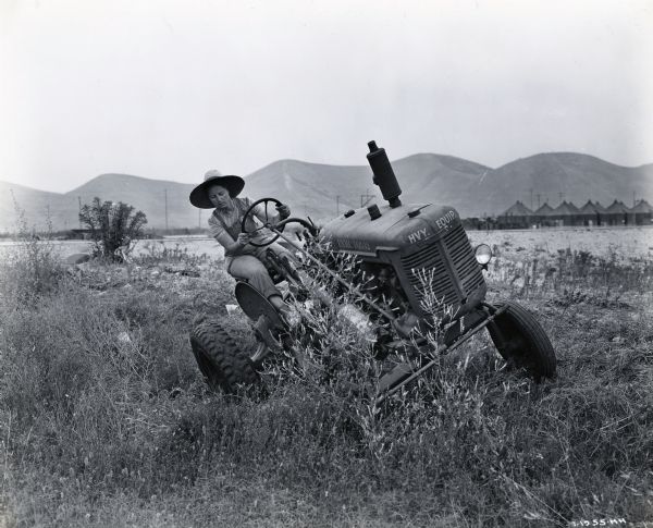 A woman wearing a wide-brimmed hat and overalls uses an International Model A tractor to maintain an area of brush at the Camp Pendleton airfield. Tents are in the background with hills behind them. The original caption reads: "International Model A tractors are used to keep grass and weeds mowed on the Camp Pendleton airfield and women civil service operators employed by the United States Navy Department operate them."