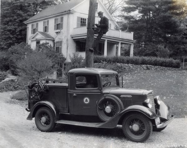 An International truck owned by Bell Systems is parked in a gravel driveway. A man wearing a hat is perched halfway up an electric pole with a coil of wire in his hands for repair. In the background is a house.