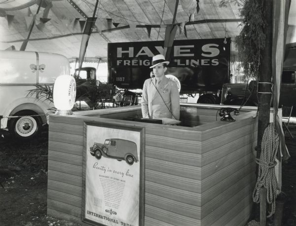 A man stands behind a booth in a tent at the Indiana State Fair. The booth is outfitted with a microphone and telephone. A poster on the front of the booth advertises International Trucks with text that reads: "Beauty in Every Line, Economy in Every Mile." A banner hanging behind the man reads: "Hayes Freight Lines."