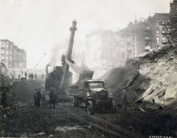 Men stand near an excavator or steam shovel as it loads an International dump truck at a construction site. The site is surrounded with multi-storied buildings. The men may be working on the New York City subway system.