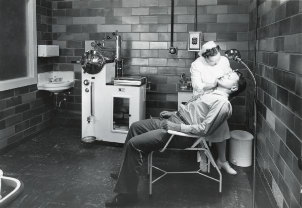 A man sits in a chair while a nurse examines his eye beneath a light in the Sterilizing and Eye Treatment Room of International Harvester's Quad Cities Tank Arsenal. Medical equipment and a sink are in the background.