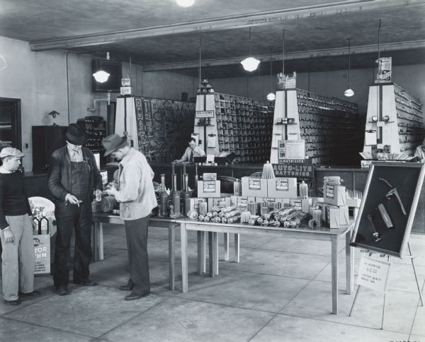 Three men stand near a display of spare parts on the sales floor of an International Harvester dealership. Another man stands behind the parts counter.